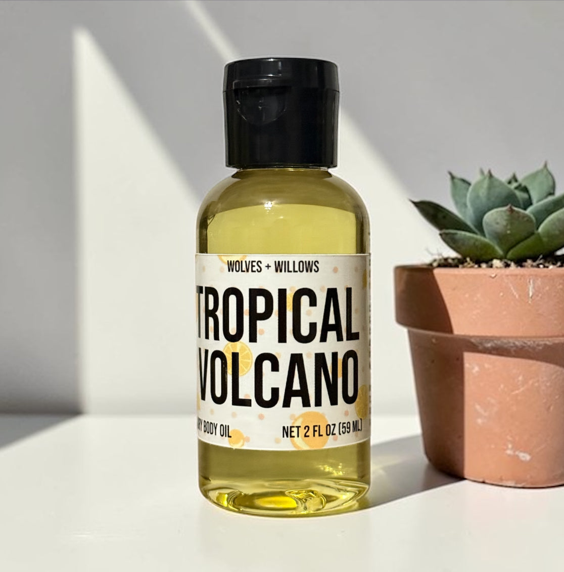 Bottle of 'Tropical Volcano' body oil by Wolves + Willows on a light surface with a potted plant in the background.