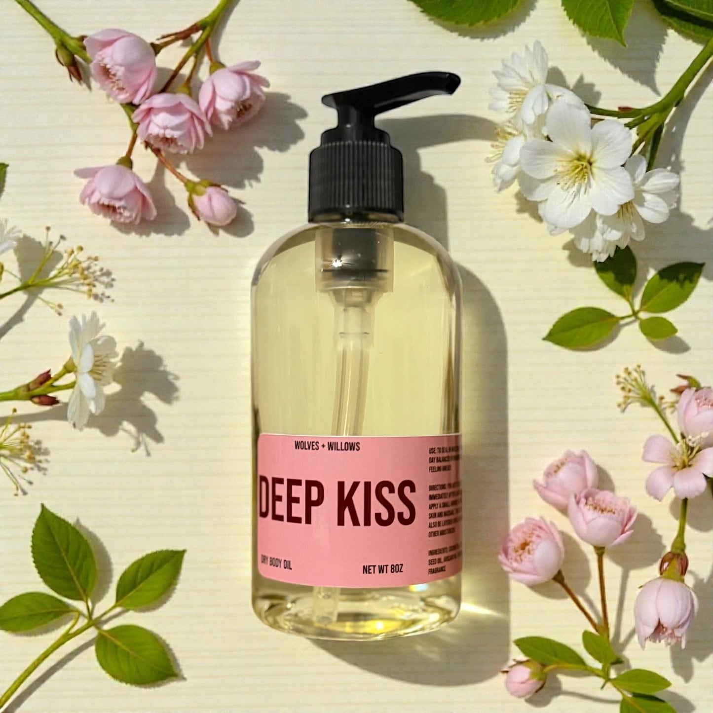 Bottle of 'Deep Kiss' body oil with black pump surrounded by flowers on a light background
