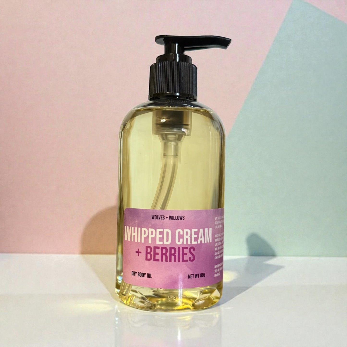 Bottle of whipped cream and berries scented dry body oil with black pump with pink label on a white surface against a pastel background.