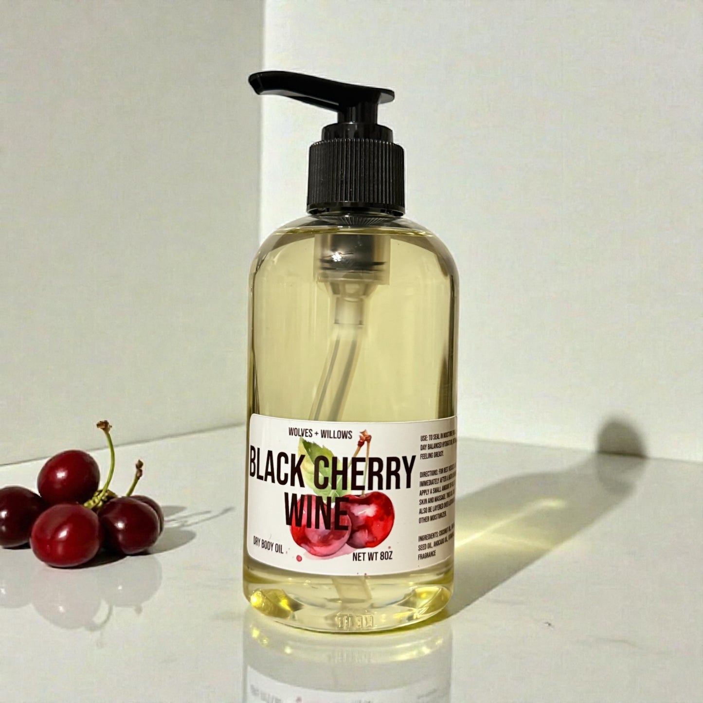 Bottle of 'Black Cherry Wine' body oil with a black pump on a white surface, surrounded by cherries.