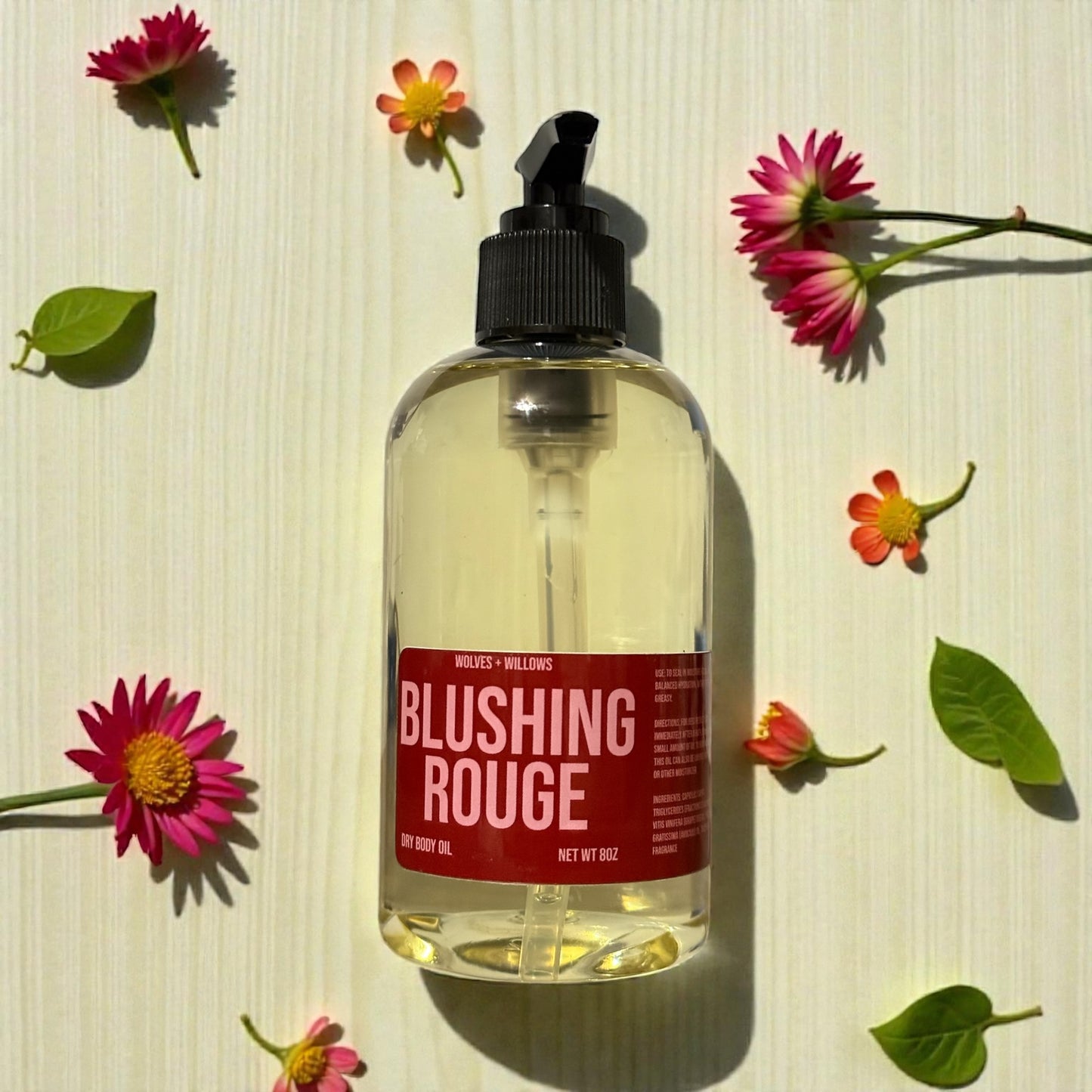 Bottle of 'Blushing Rouge' body oil with a red label on a light wooden surface surrounded by flowers and leaves.
