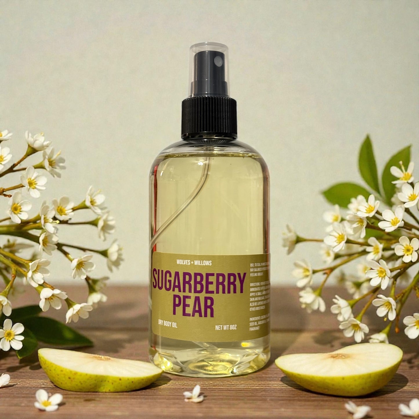 Bottle of 'Sugarberry Pear' dry body oil with flowers and an apple on a wooden surface.