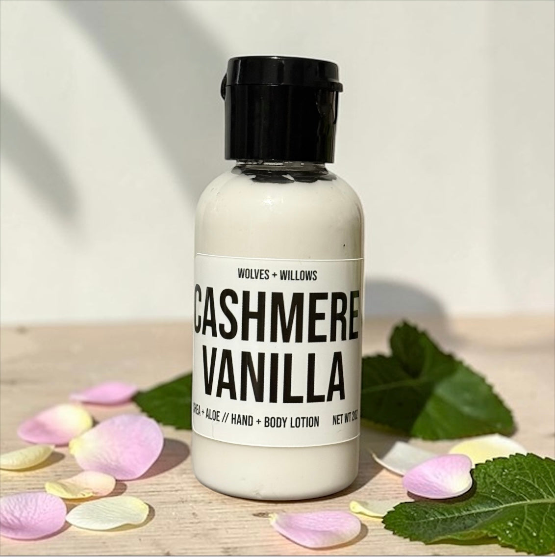 Bottle of 'Cashmere Vanilla' lotion with a black cap on a wooden surface with leaves and petals.