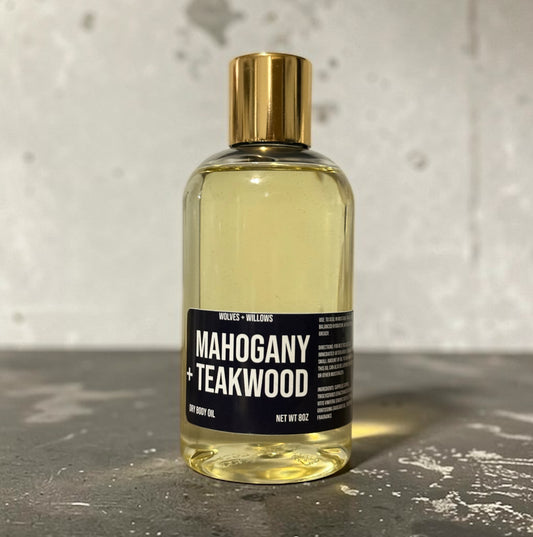 Bottle of Mahogany + Teakwood body oil with a gold cap on a textured surface.