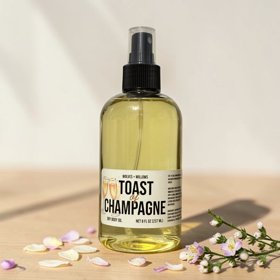 Bottle of 'Toast of Champagne' dry body oil with a label on a wooden surface with flowers and petals.