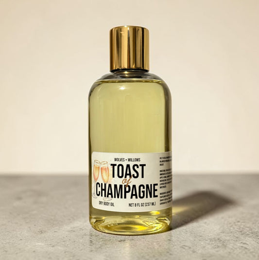 Bottle of 'Toast of Champagne' body oil with a gold cap on a neutral background