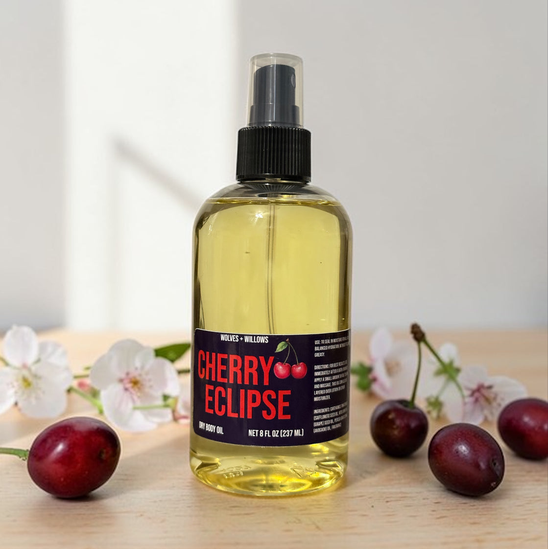 Bottle of 'Cherry Eclipse' body oil with cherries and flowers on a wooden surface