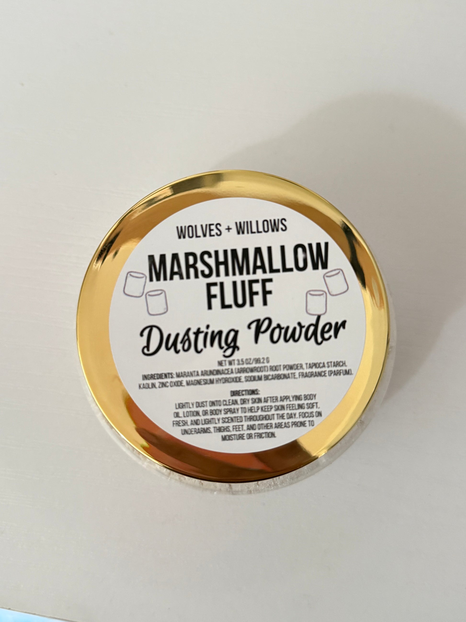 A round tin of Wolves + Willows Marshmallow Fluff Dusting Powder, featuring a shiny gold metallic rim and a white label with black text, photographed on a plain off-white surface.