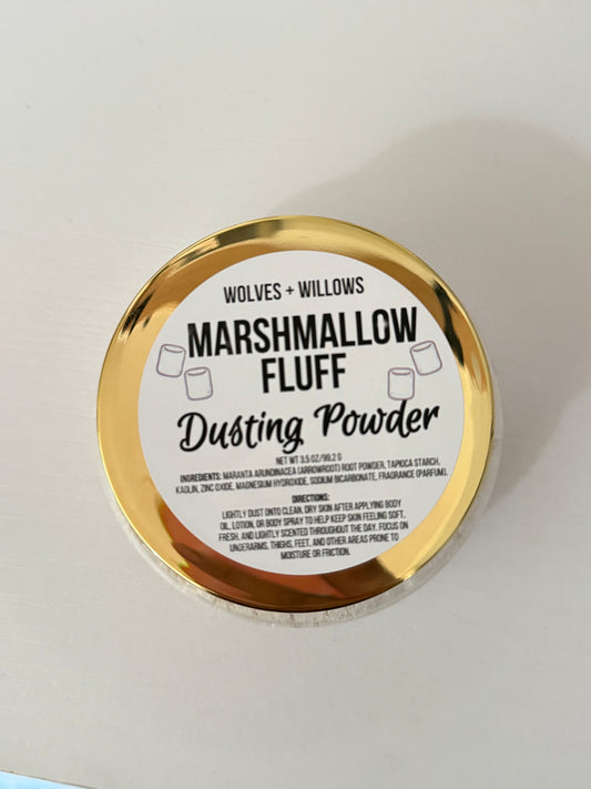 A round tin of Wolves + Willows Marshmallow Fluff Dusting Powder, featuring a shiny gold metallic rim and a white label with black text, photographed on a plain off-white surface.
