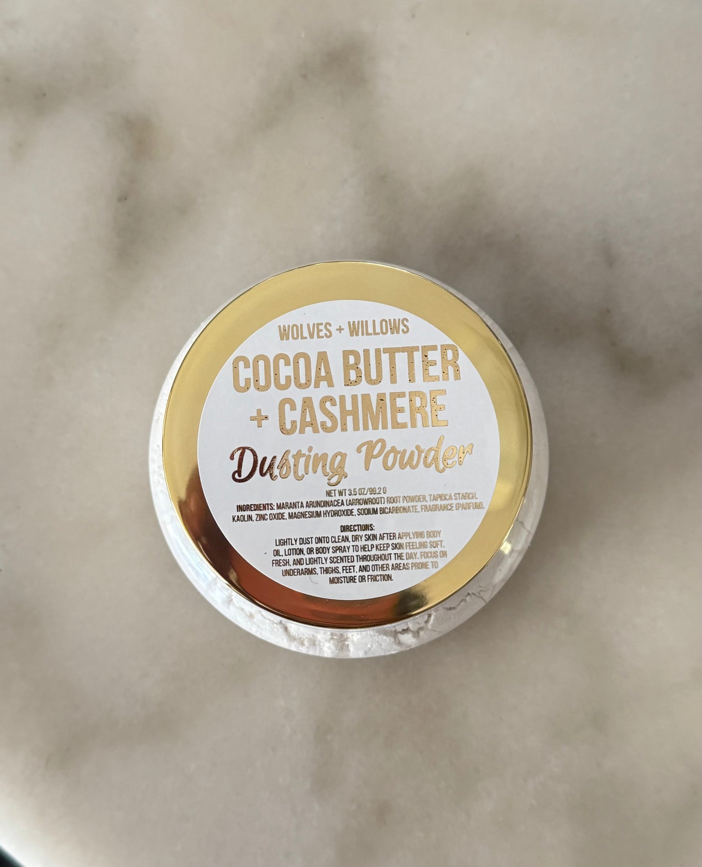 A round tin of Wolves + Willows Cocoa Butter + Cashmere Dusting Powder, viewed from above on a light marble surface. The lid features a gold and white label with gold lettering.