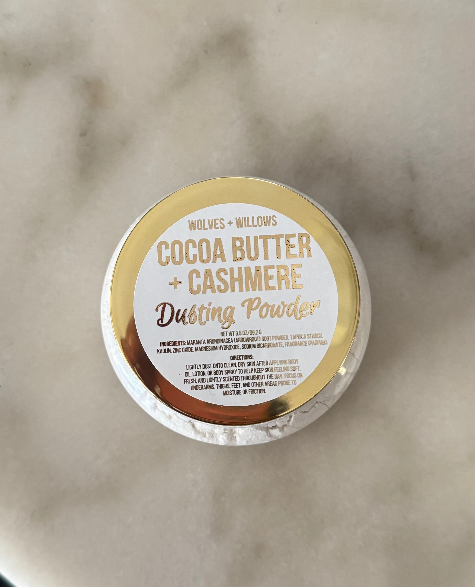A round tin of Wolves + Willows Cocoa Butter + Cashmere Dusting Powder, viewed from above on a light marble surface. The lid features a gold and white label with gold lettering.
