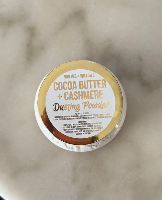A round tin of Wolves + Willows Cocoa Butter + Cashmere Dusting Powder, viewed from above on a light marble surface. The lid features a gold and white label with gold lettering.