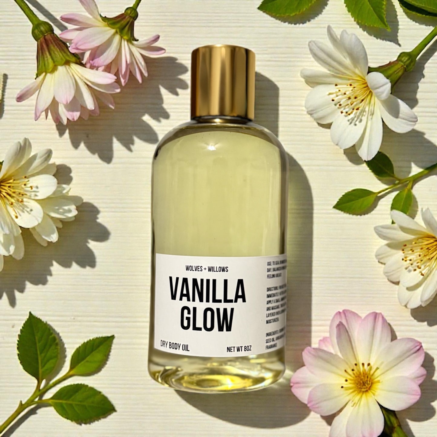Bottle of Vanilla Glow body oil surrounded by flowers on a light background