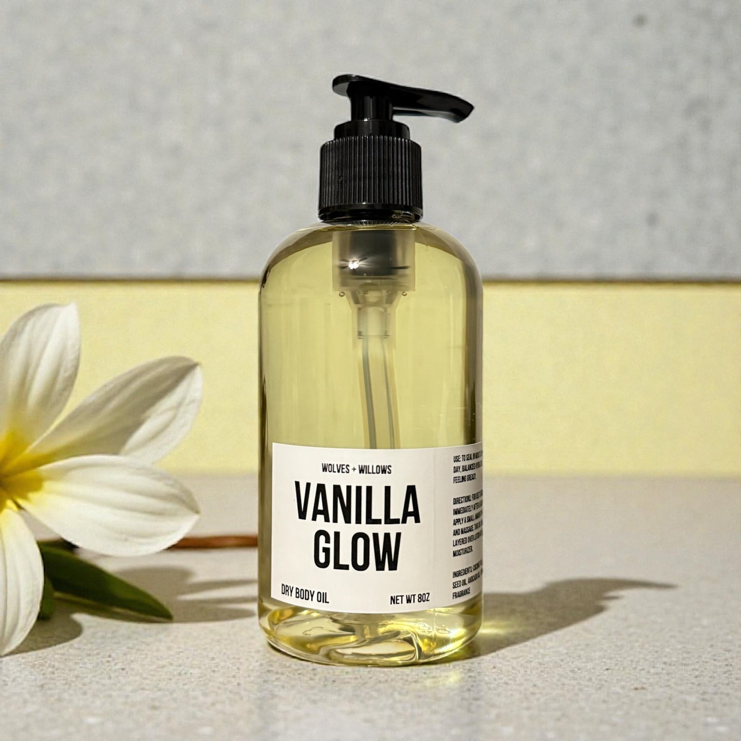 Bottle of 'Vanilla Glow' dry body oil with a white flower on a light surface.