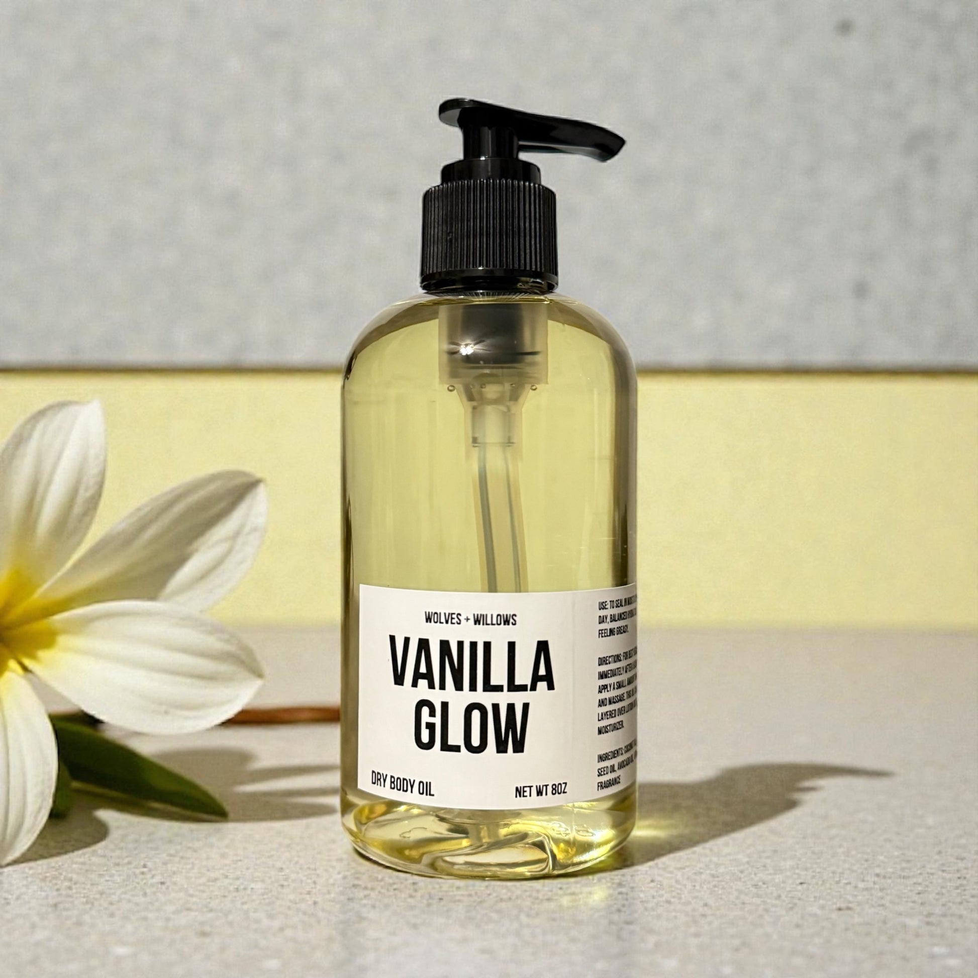 Bottle of 'Vanilla Glow' dry body oil with a white flower on a light surface.