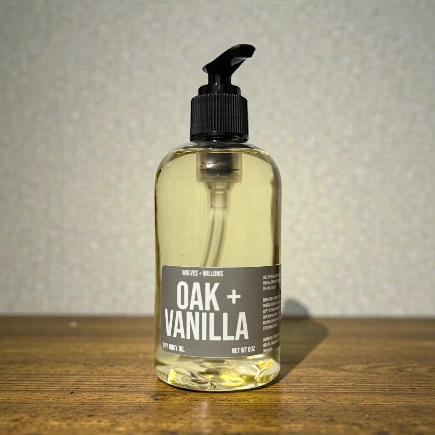 8 ounce bottle of  'Oak + Vanilla' dry body oil, with black pump, on a wooden surface with a neutral background
