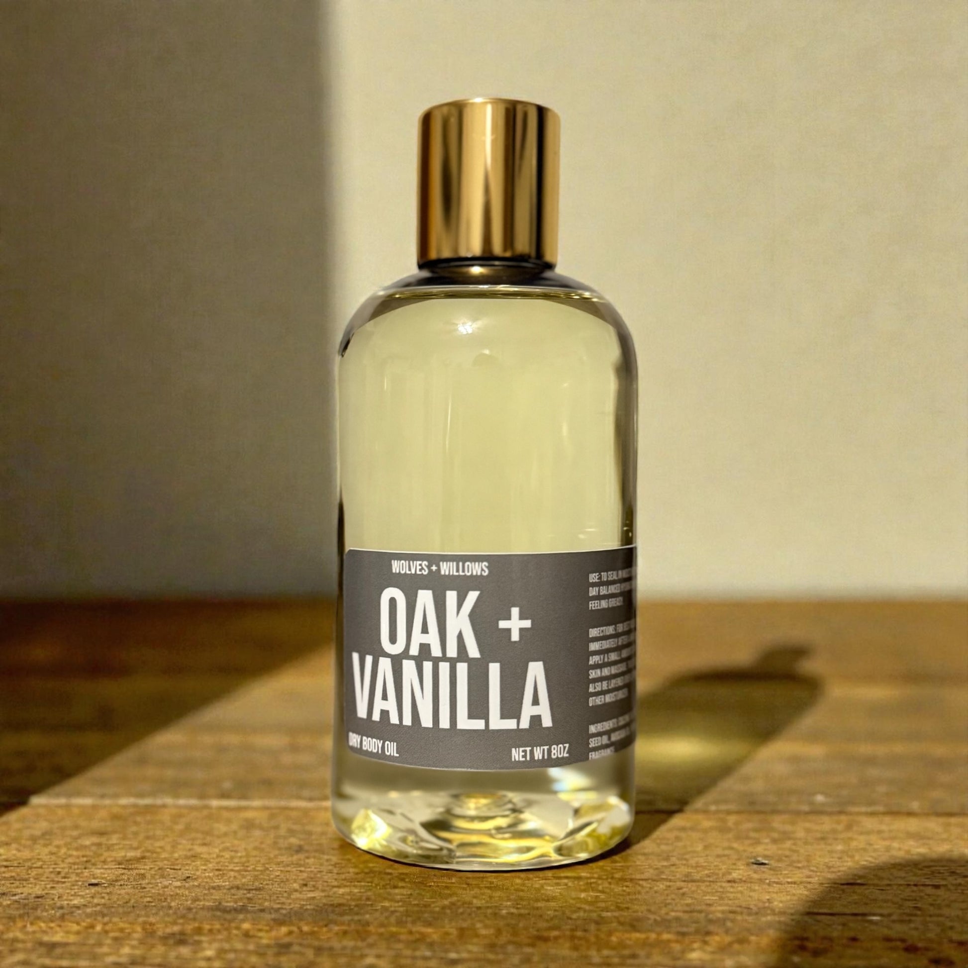 8 ounce bottle of  'Oak + Vanilla' body oil, with gold dispensing cap, on a wooden surface with a neutral background
