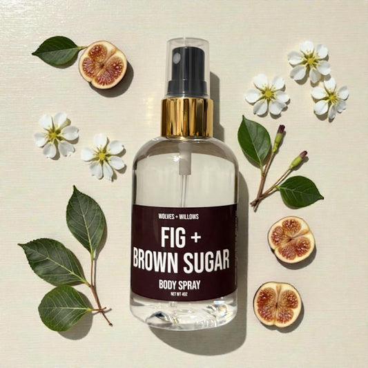 Body spray bottle labeled 'Fig + Brown Sugar' surrounded by figs and flowers on a beige background