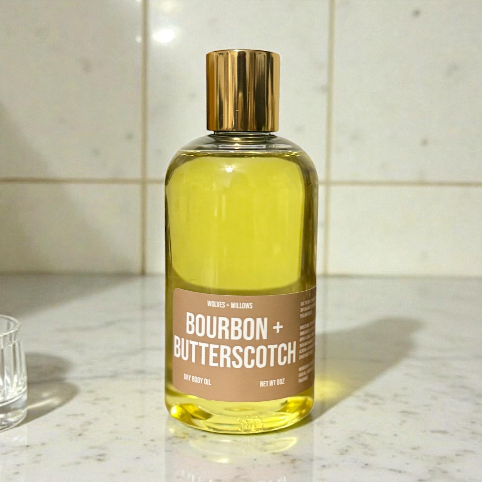 8 ounce "bourbon + butterscotch" body oil in clear bottle with gold dispensing cap.