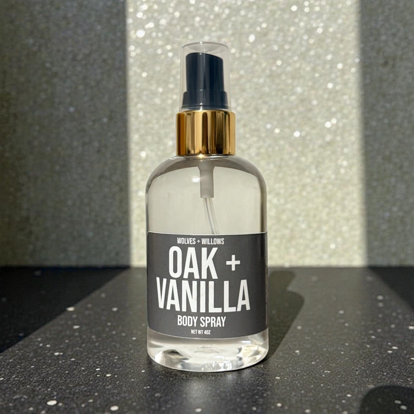 Body spray bottle labeled 'Oak + Vanilla' on a reflective surface with a glittery background.