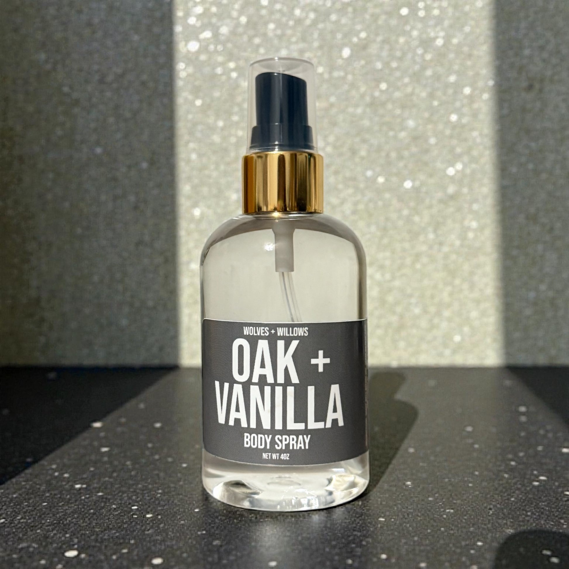 Body spray bottle labeled 'Oak + Vanilla' on a reflective surface with a glittery background.