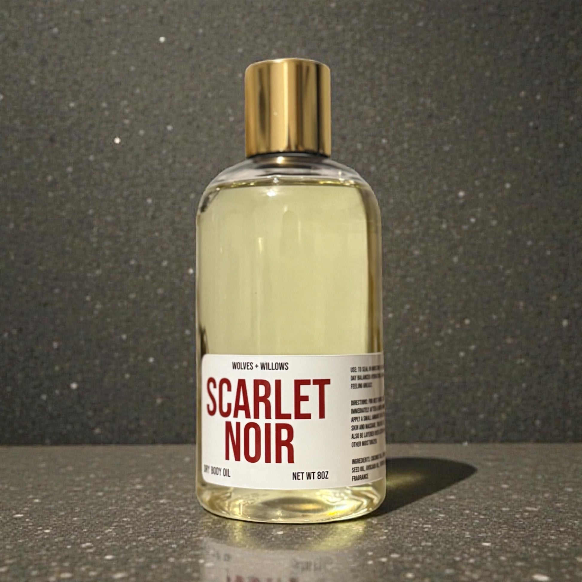 Bottle of 'Scarlet Noir' body oil with a gold cap on a reflective surface.
