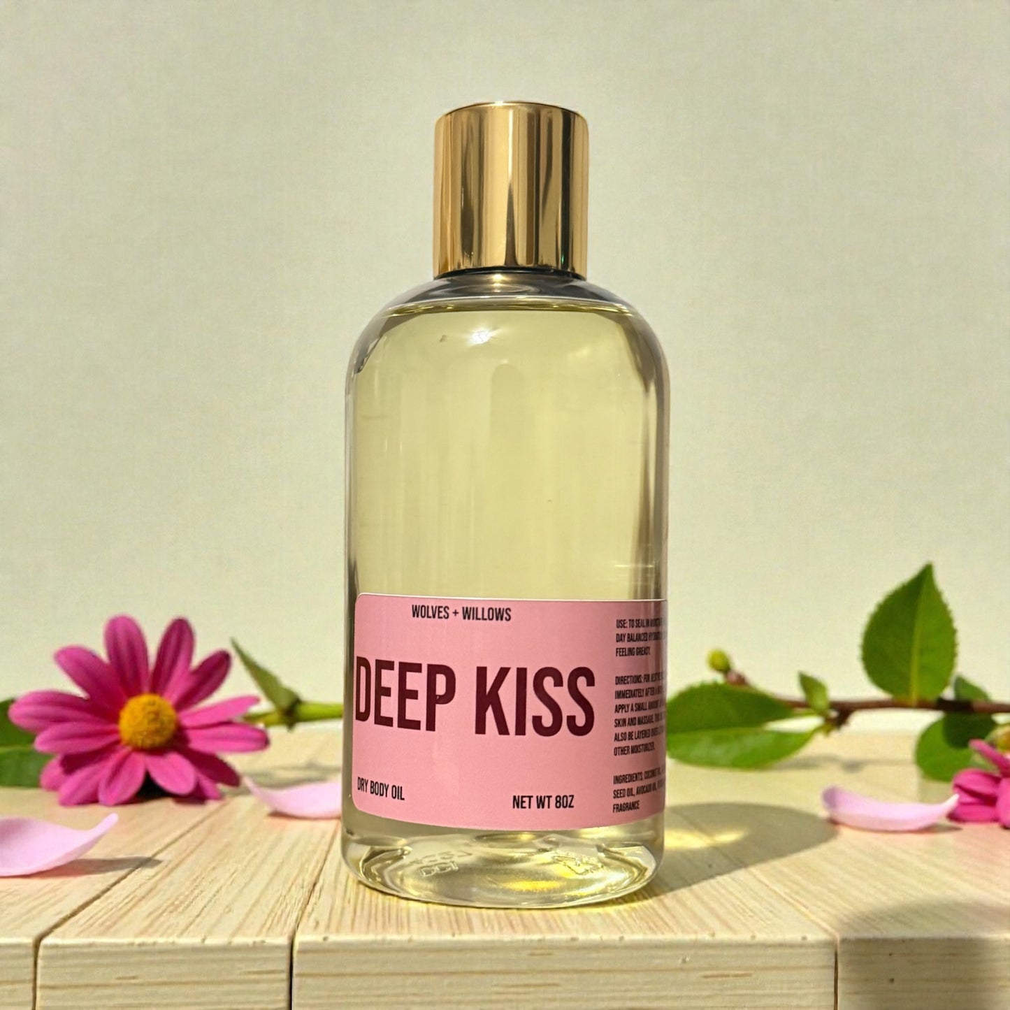 Bottle of 'Deep Kiss' body oil with gold pour cap on a wooden surface with flowers.