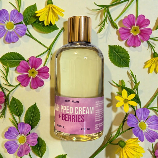 Bottle of 'Whipped Cream + Berries' body oil with gold pour cap surrounded by flowers on a light background