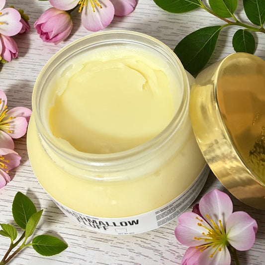 Jar of "Marshmallow Fluff" creamy body butter with a gold lid on a wooden surface with flowers and leaves.