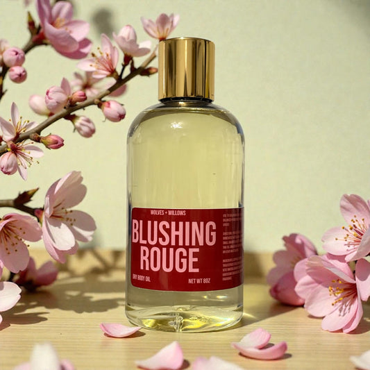 Bottle of 'Blushing Rouge' body oil with pink flowers on a wooden surface