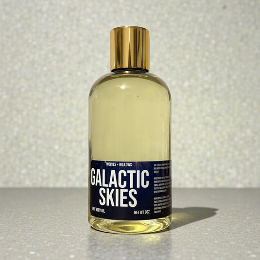 Bottle of 'Galactic Skies' dry body oil with a gold cap on a reflective surface.