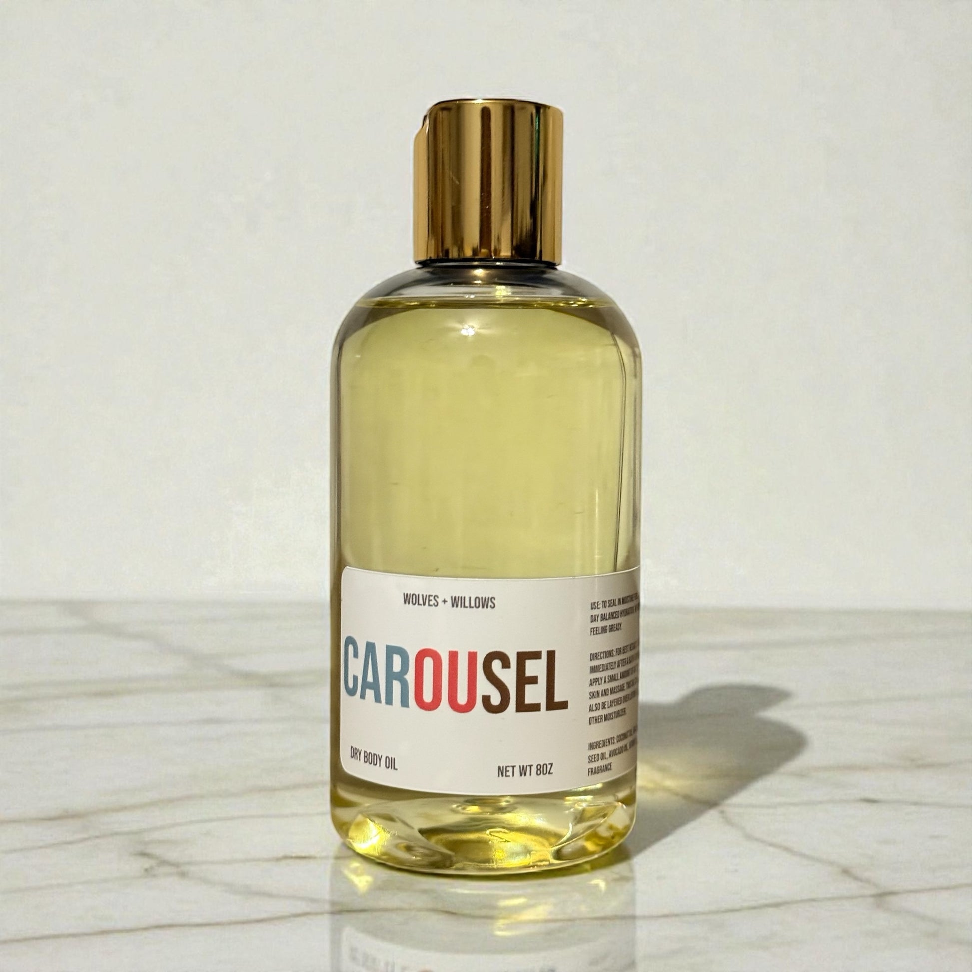Bottle of 'Carousel' dry body oil with a gold cap on a marble surface.