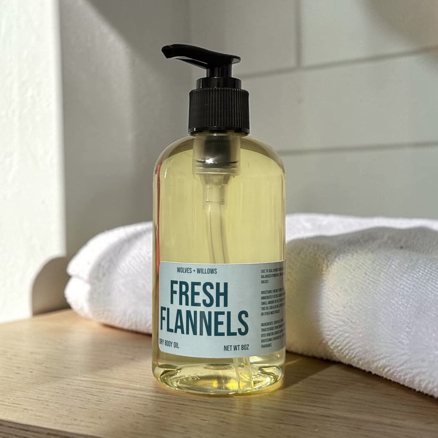 Bottle of 'Fresh Flannels' dry body oil on a wooden surface with a white towel in the background.
