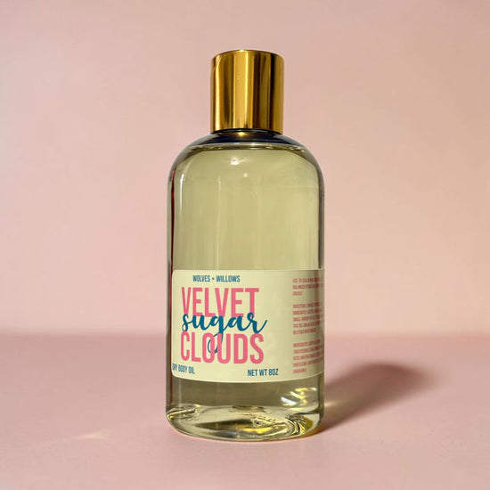 Bottle of 'Velvet Sugar Clouds' body oil with a gold cap on a pink background