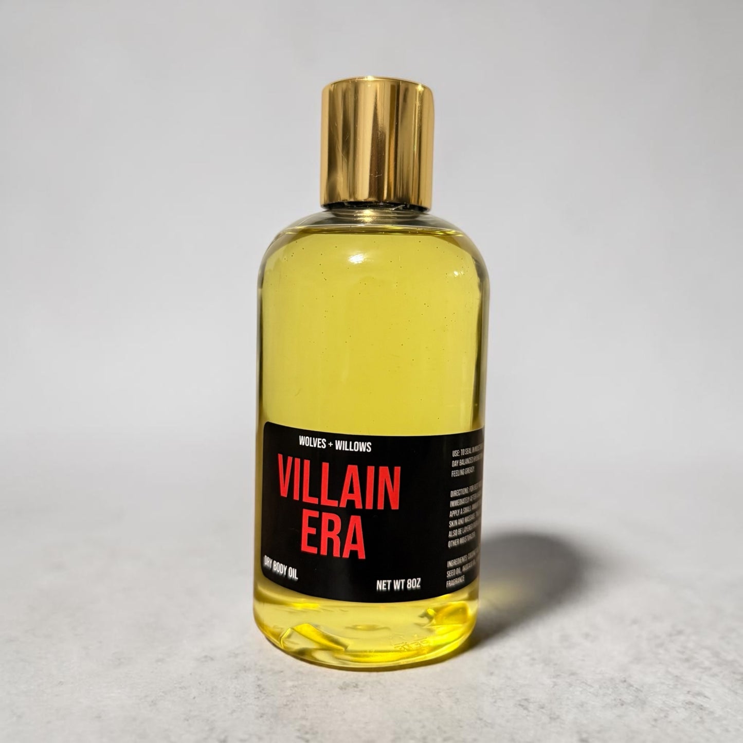 Bottle of 'Villain Era' body oil with a gold cap on a light gray background