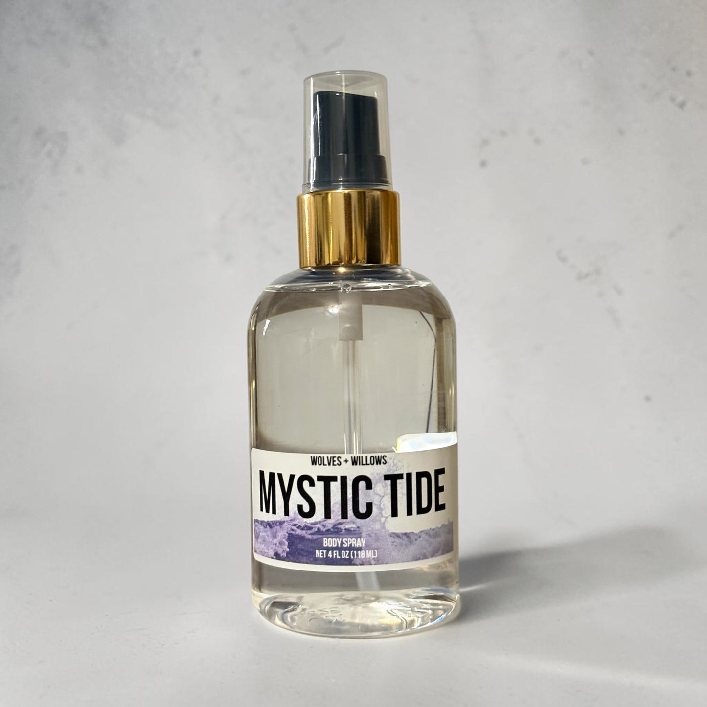 A clear glass spray bottle of Wolves + Willows 'Mystic Tide' hand spray with a gold collar and black sprayer, set against a light gray textured background.