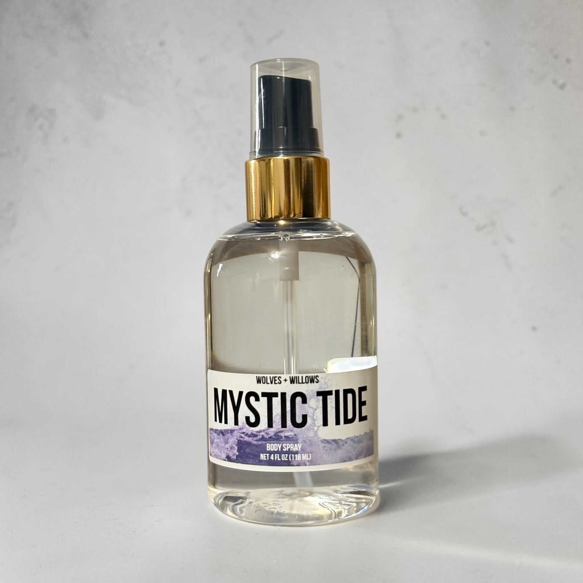 A clear glass spray bottle of Wolves + Willows 'Mystic Tide' hand spray with a gold collar and black sprayer, set against a light gray textured background.