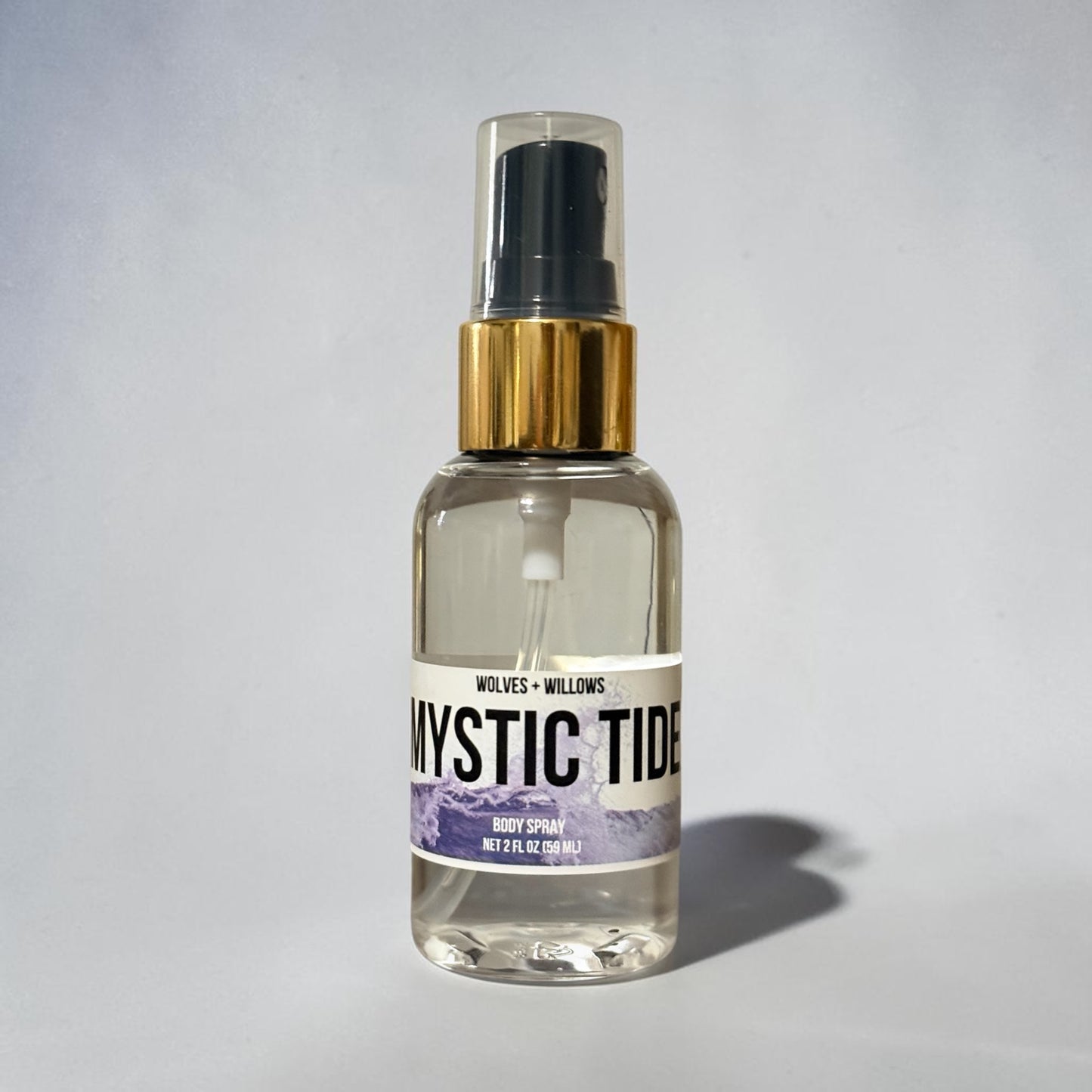 A clear glass body spray bottle with a black spray top, gold collar, and a white label reading 'Mystic Tide' with purple mountain artwork. The bottle is centered on a light gray background with a soft shadow to the right.