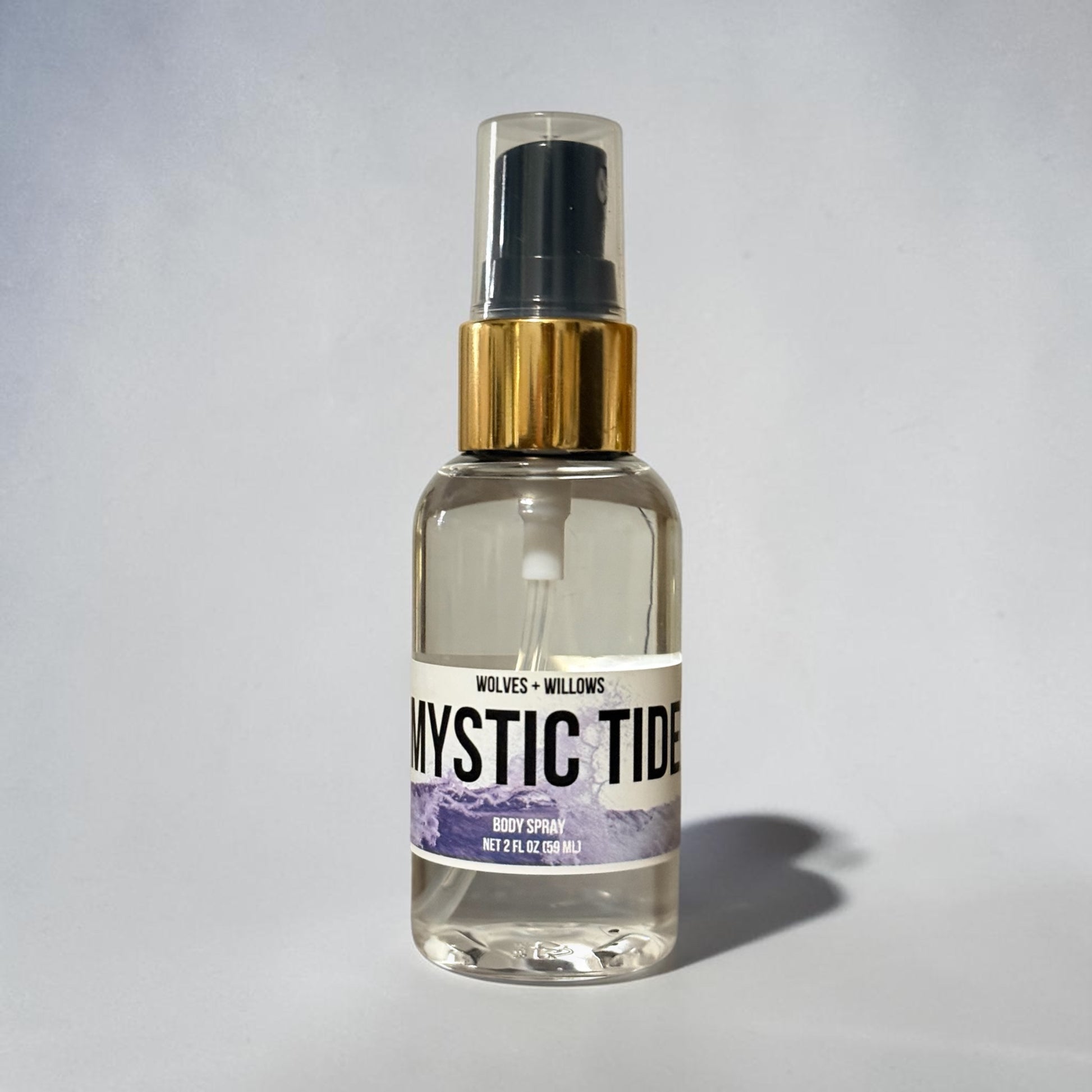 A clear glass body spray bottle with a black spray top, gold collar, and a white label reading 'Mystic Tide' with purple mountain artwork. The bottle is centered on a light gray background with a soft shadow to the right.