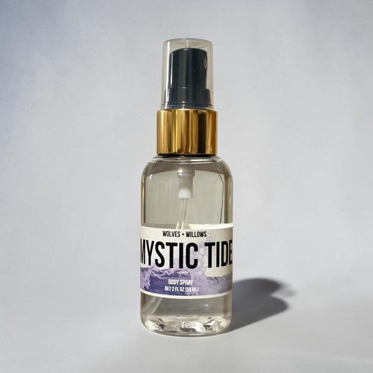 A clear glass body spray bottle with a black spray top, gold collar, and a white label reading 'Mystic Tide' with purple mountain artwork. The bottle is centered on a light gray background with a soft shadow to the right.