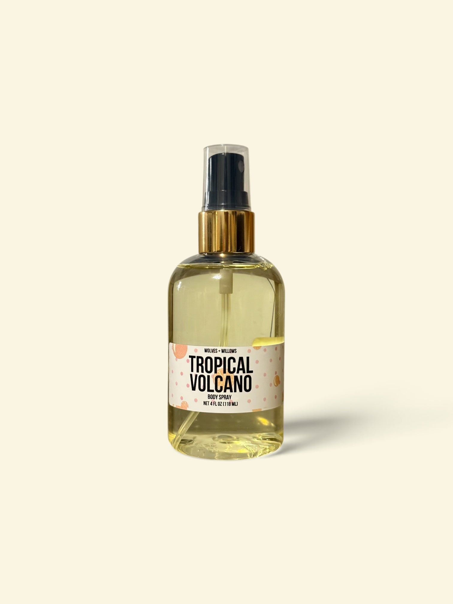 A clear spray bottle filled with pale yellow liquid, featuring a black spray top, gold collar, and a cream label that reads 'TROPICAL VOLCANO'. The bottle is centered against a solid cream background with a soft shadow.