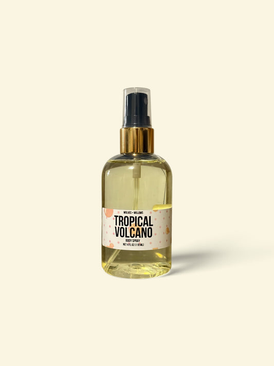 A clear spray bottle filled with pale yellow liquid, featuring a black spray top, gold collar, and a cream label that reads 'TROPICAL VOLCANO'. The bottle is centered against a solid cream background with a soft shadow.