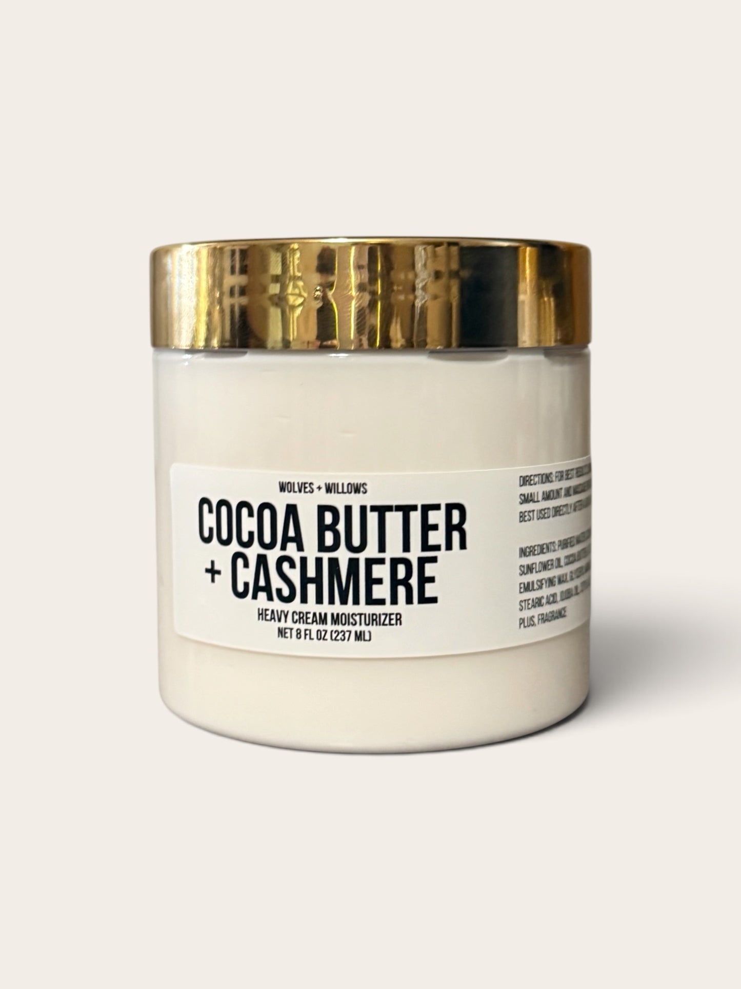 A jar of Wolves + Wills Cocoa Butter + Cashmere heavy cream moisturizer, featuring an off-white jar and a shiny gold metallic lid, set against a solid light beige background.