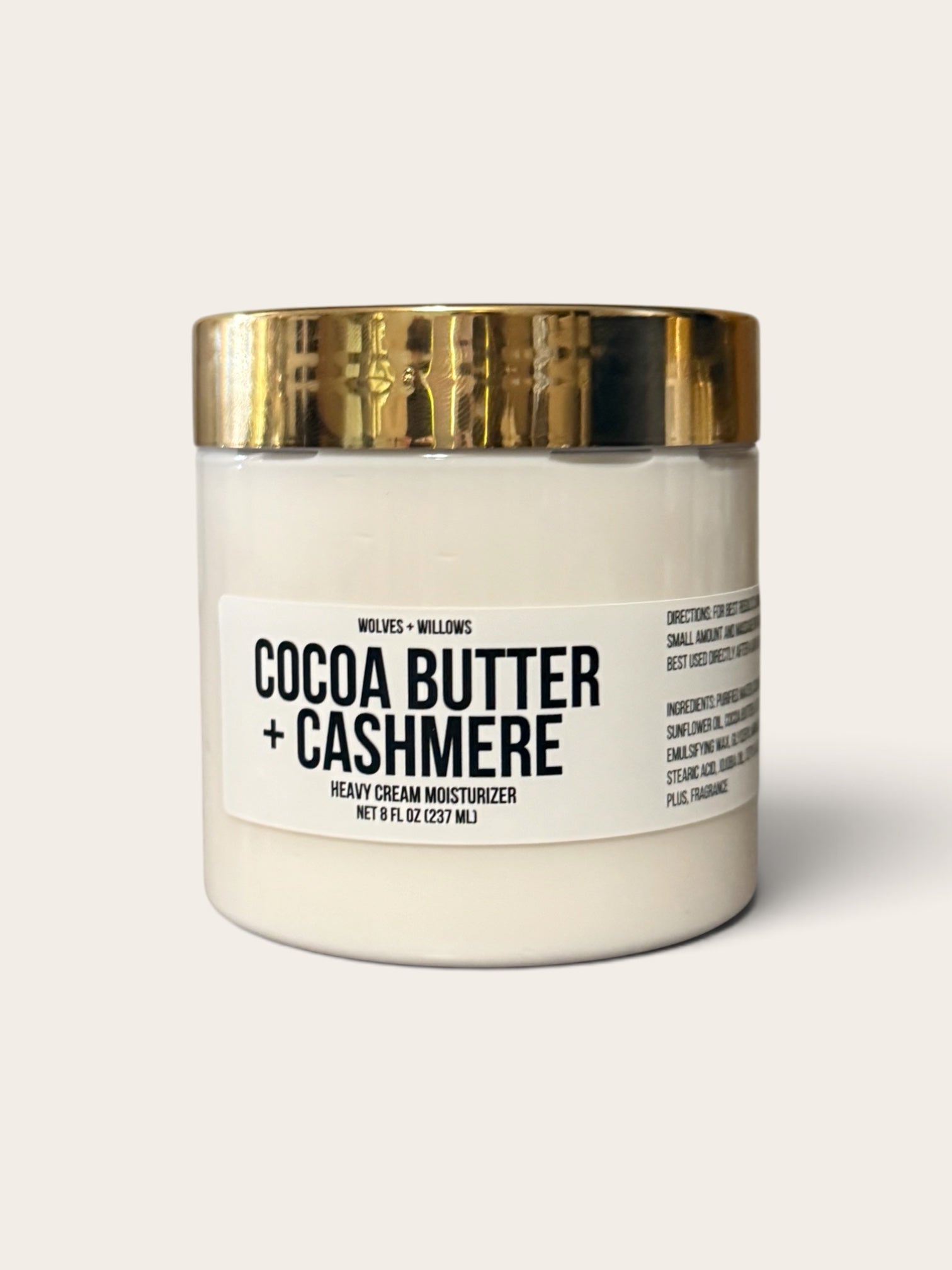 A jar of Wolves + Wills Cocoa Butter + Cashmere heavy cream moisturizer, featuring an off-white jar and a shiny gold metallic lid, set against a solid light beige background.
