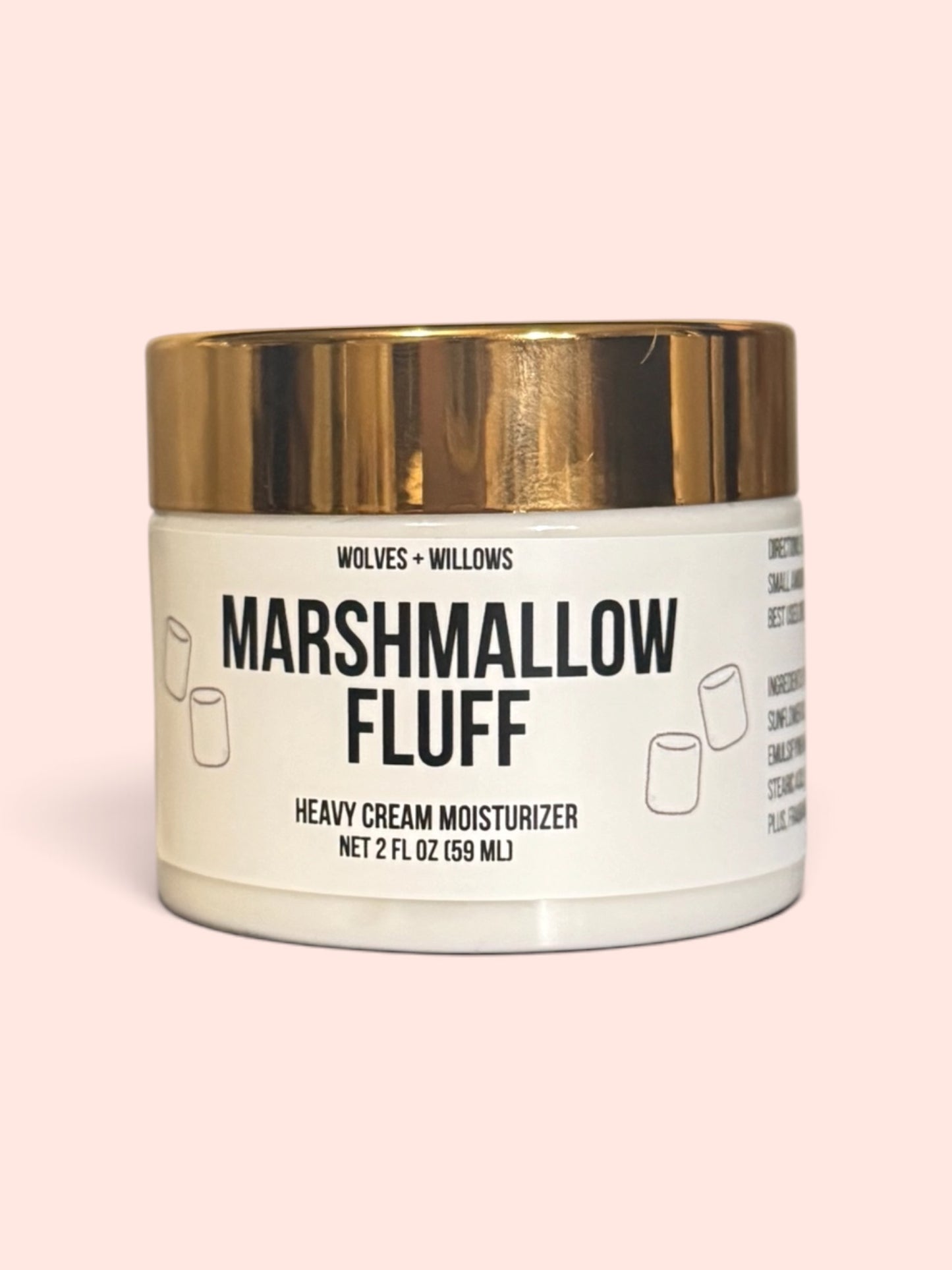 A jar of Wolves + Willows Marshmallow Fluff Heavy Cream Moisturizer, featuring a white container with a metallic gold lid and a white label with black text, set against a solid pale pink background.