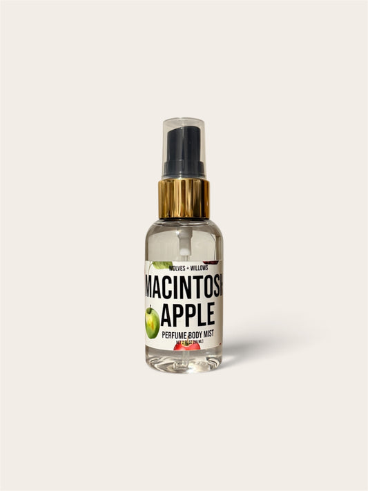 A clear glass perfume body mist bottle with a black spray top, gold collar, and a white label reading 'MACINTOSH APPLE' with apple graphics, centered on a light warm gray background.