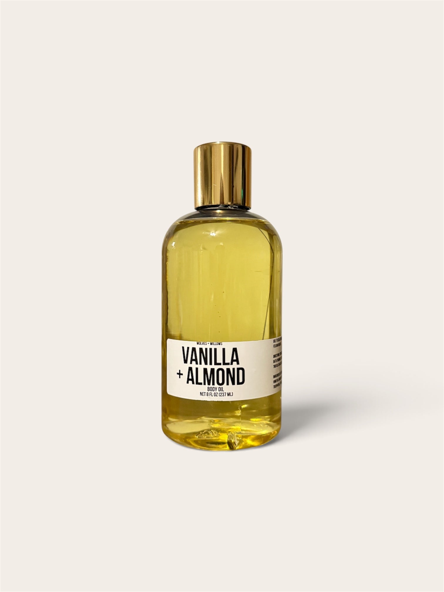 A clear bottle filled with pale yellow liquid, featuring a metallic gold cap and a white label that reads 'VANILLA + ALMOND'. The bottle is centered against a solid, warm off-white background.