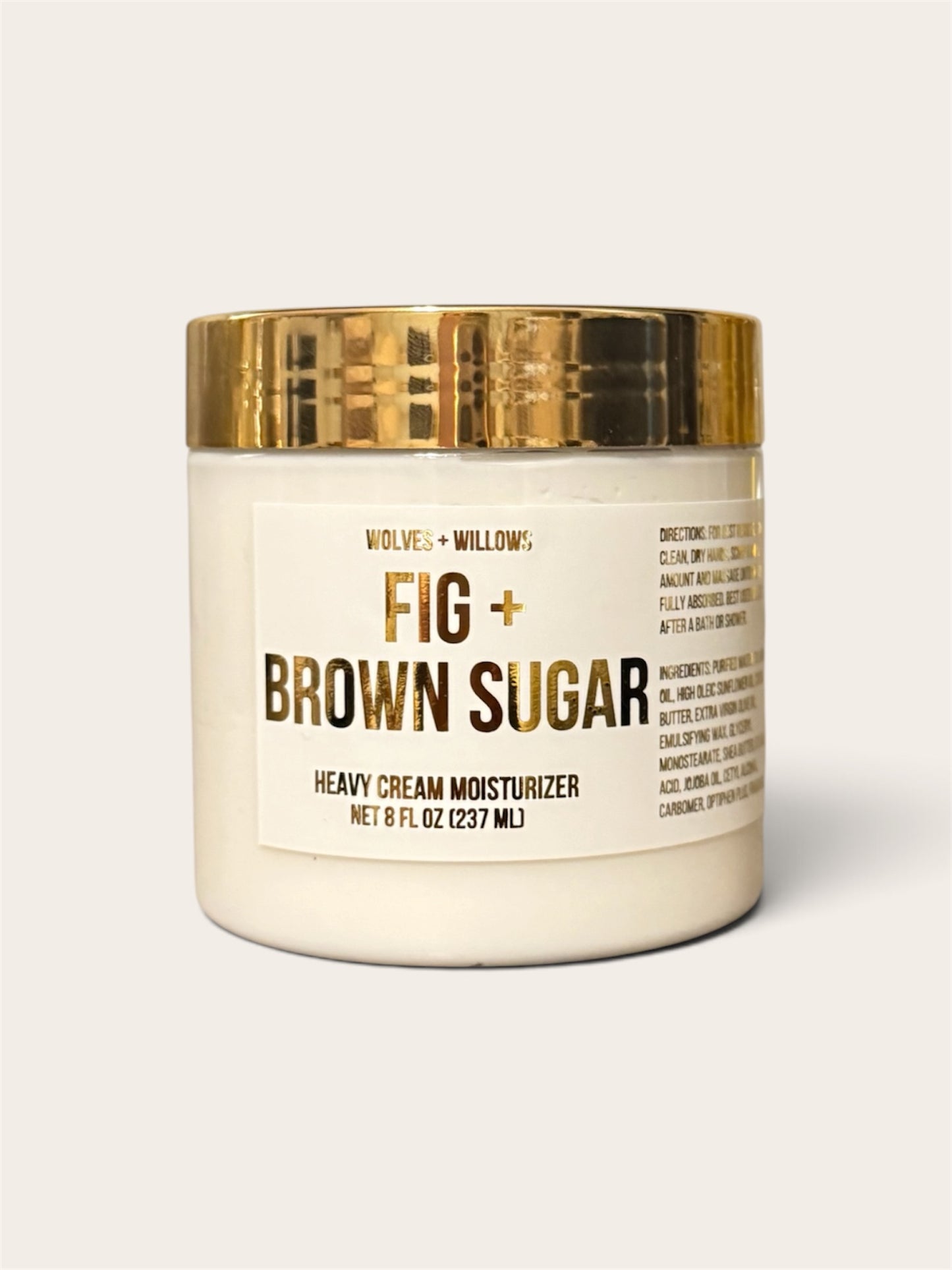A jar of Wolves + Willows Fig + Brown Sugar Heavy Cream Moisturizer, featuring an off-white body, a textured metallic gold lid, and a cream-colored label with gold and black text, set against a solid light beige background.