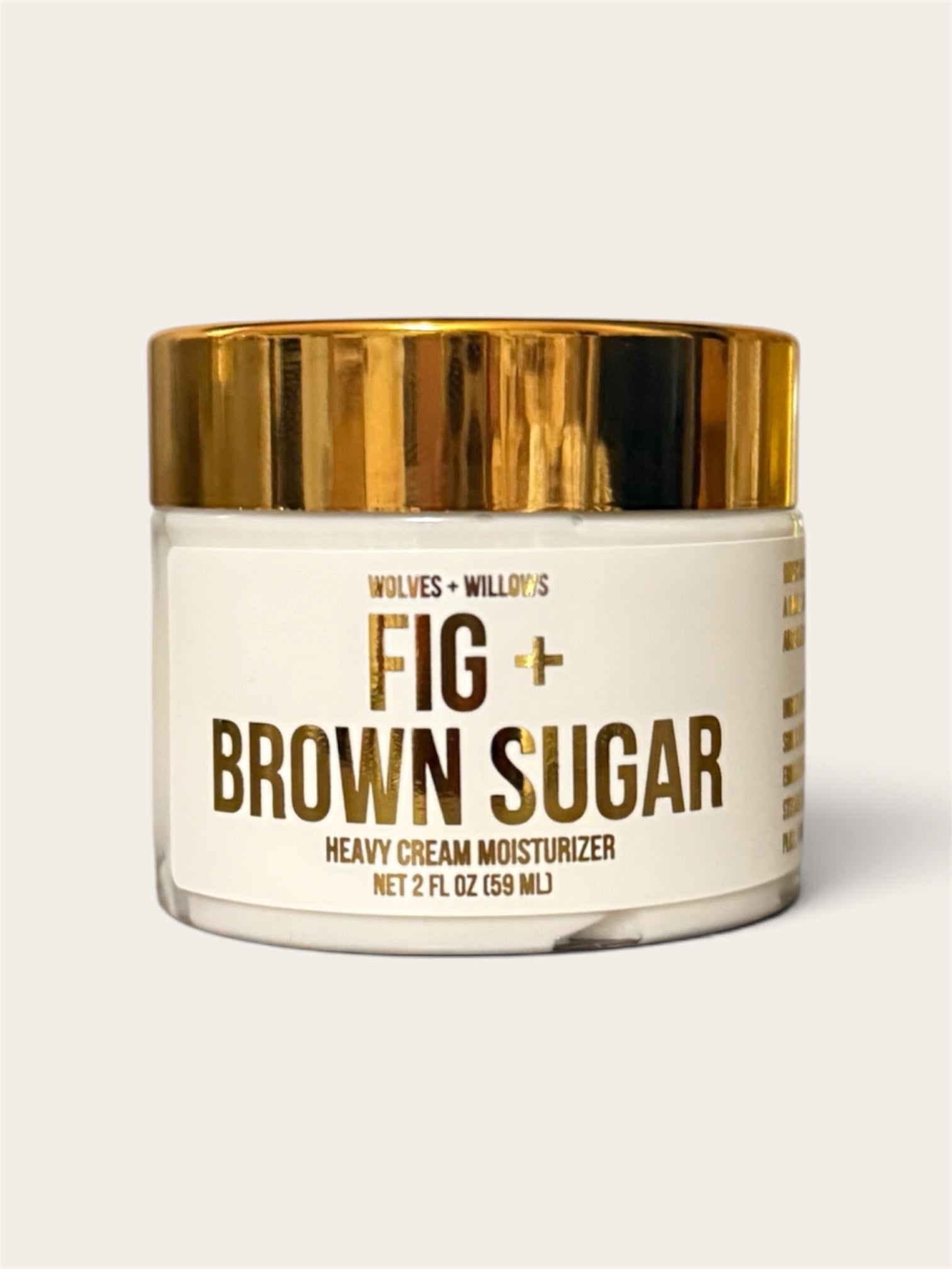 A jar of Wolves + Willow's Fig + Brown Sugar Heavy Cream Moisturizer, featuring a white label and a shiny gold metallic lid, set against a solid light beige background.